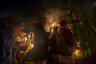 A group of Maya farmers gathered around a smoky fire at night, holding lit candles in a dimly lit outdoor setting with plants and trees in the background.